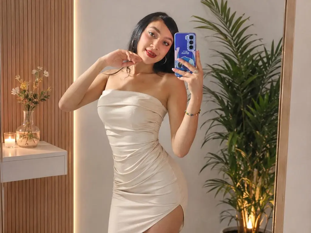 EvelynClaiir poses in a tight, strapless cream-colored dress, holding a blue phone for a mirror selfie in a room with a plant and warm lighting.