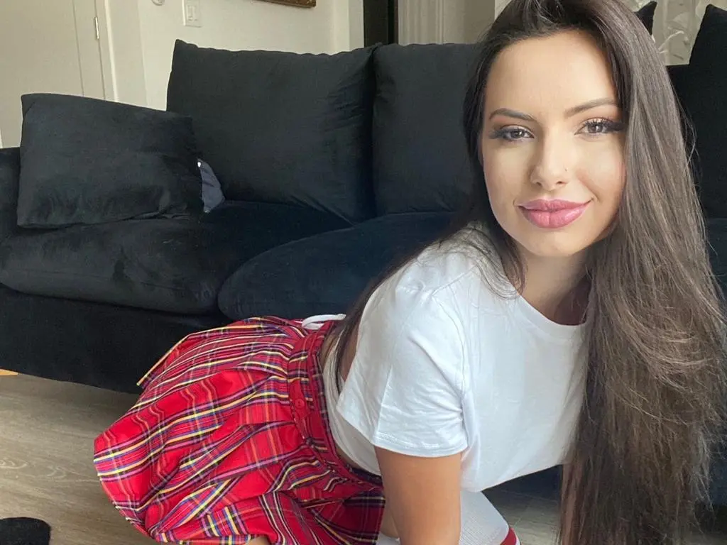 Stassia smiles while wearing a white t-shirt and a red plaid skirt, sitting on the floor in front of a black couch.