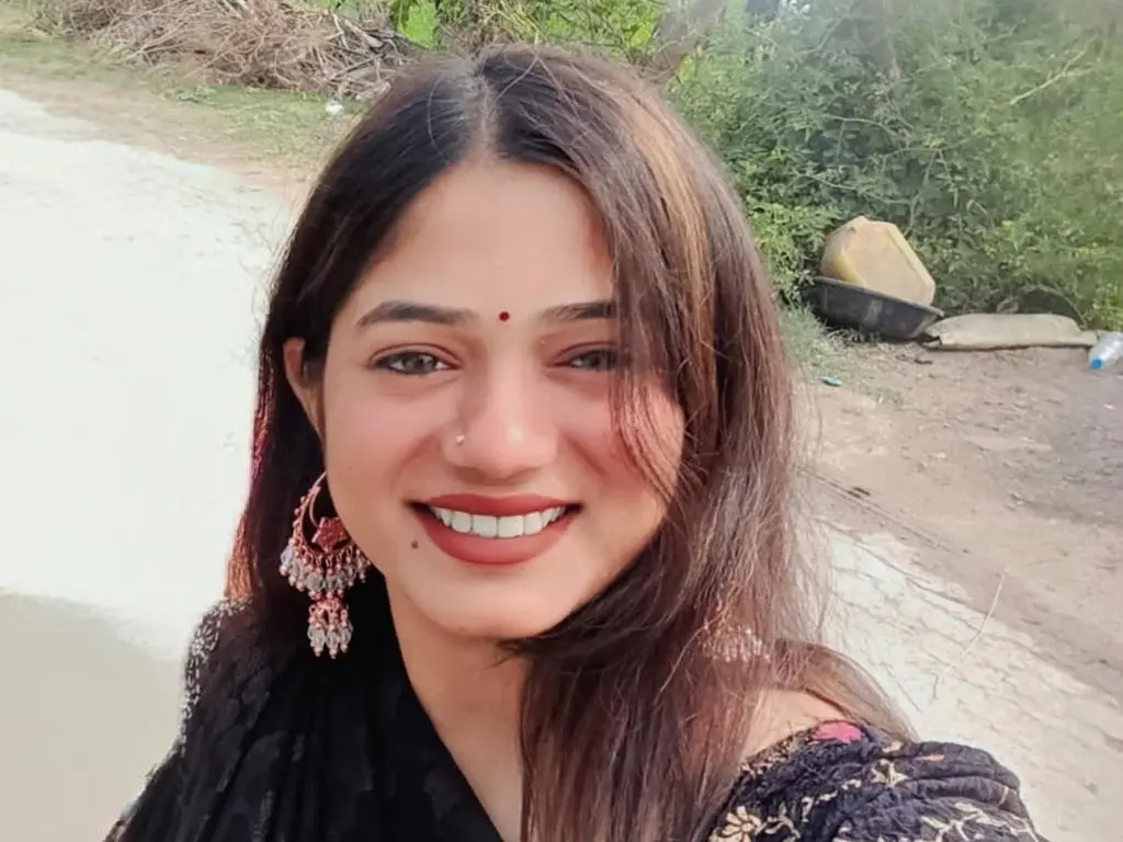 Bmw_black smiles while wearing large pink earrings and a black patterned top, with long brown hair and a red bindi on her forehead.