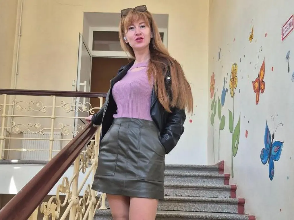 AnissaDolll is a long-haired woman wearing a purple top and a black leather skirt, posing on a staircase next to a floral wall mural.