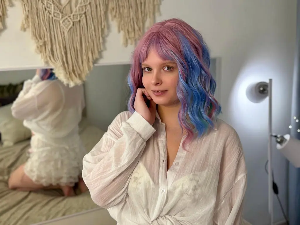 Stacy_Star poses in a white shirt and lace bra, showing off her colorful pink and blue hair while sitting on a bed.