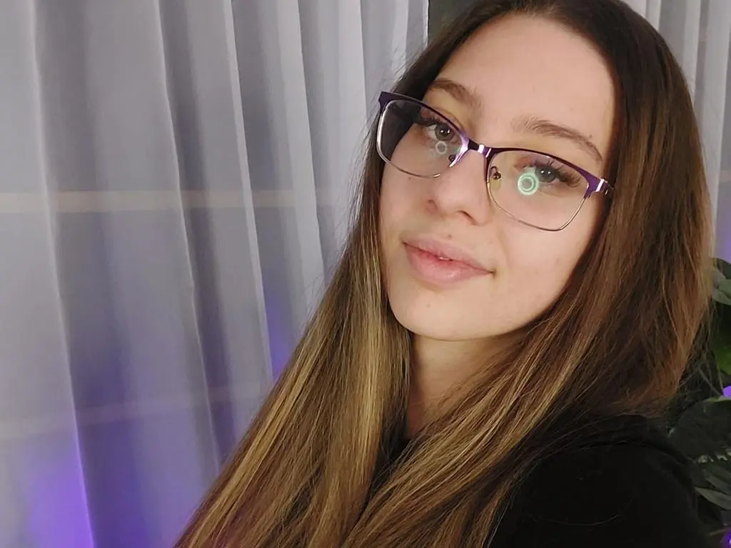 RavenBlliss wearing purple glasses and a black top, posing with long brown hair against a light curtain background.