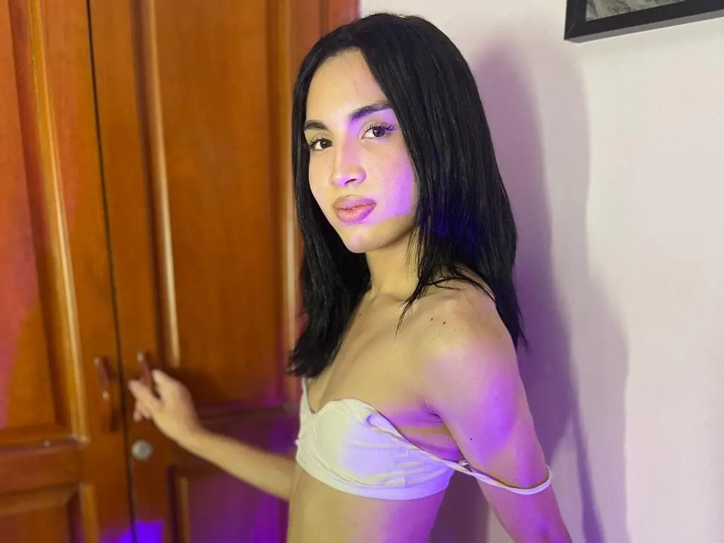 JackyMurphy1 with long black hair poses in a white bra against a wooden door and white wall.