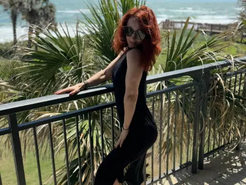 Redheaded woman in a black jumpsuit posing on a sunny balcony overlooking tropical palm trees and the ocean.