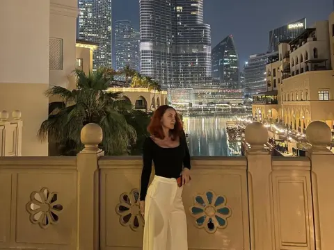 Diamond_Slutty — Elegant woman in a black top and white trousers standing on a balcony overlooking a glowing city skyline at night.