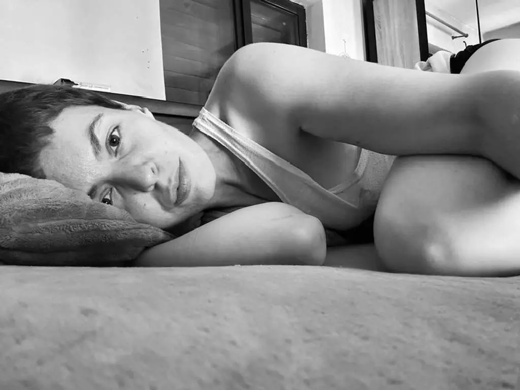 Relaxed woman in a white tank top lying on a bed in a bright, minimalist bedroom setting.