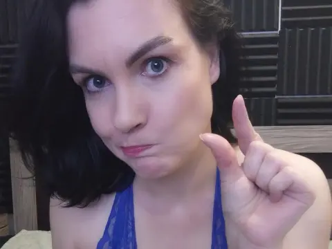 Playful young woman with dark hair in a blue lace top making a finger gun gesture in a cozy indoor setting.