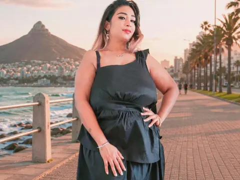princesSsandy — princesSsandy stands on a seaside boardwalk in a black sleeveless dress with the ocean and mountains in the background.
