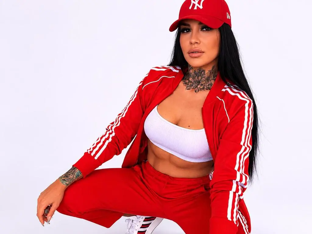 HottyNicole — Woman in red tracksuit and white crop top wearing a red baseball cap against a white background.