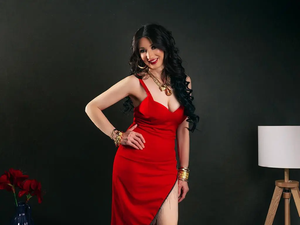 Smiling woman in a glamorous red evening gown and gold jewelry posing against a dark studio background.