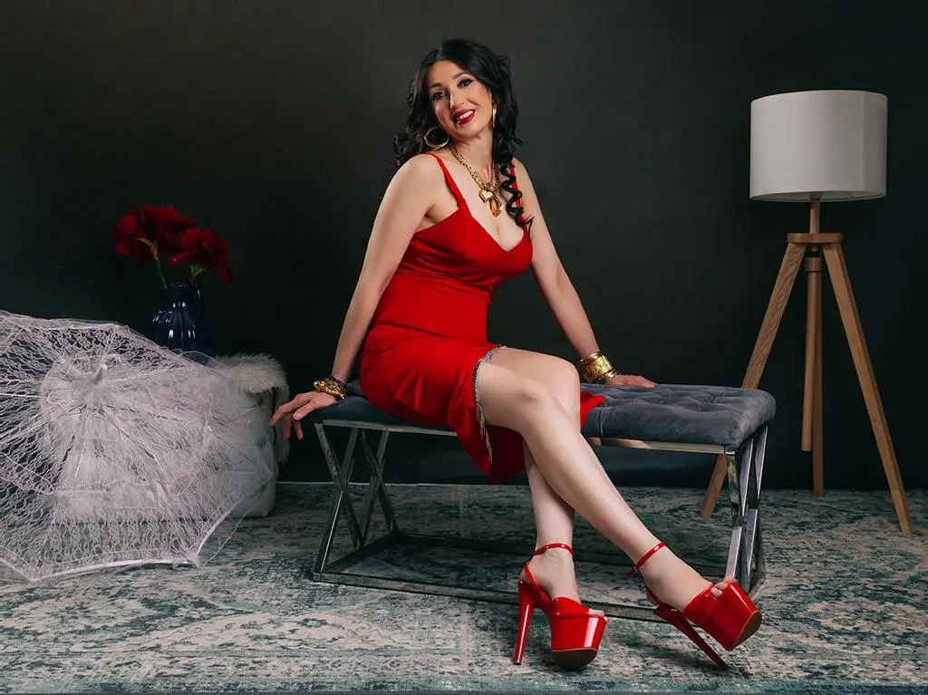 SpecialCerise — SpecialCerise sits on a grey bench wearing a red slip dress and red high heels, posing against a dark background.
