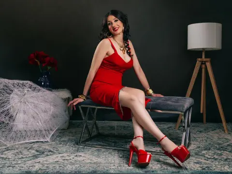 Elegant woman in a red cocktail dress sitting on a velvet bench in a moody, dark studio setting.