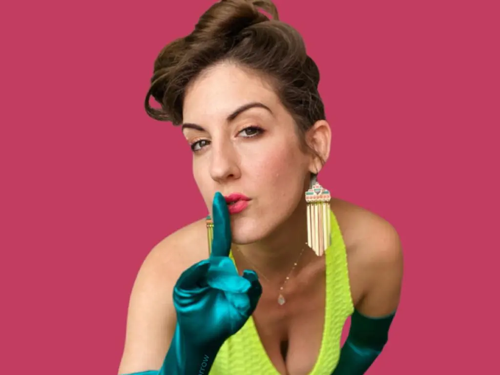 Ms_Tomorrow poses with a finger to her lips, wearing bright yellow top and long teal gloves against a pink background.