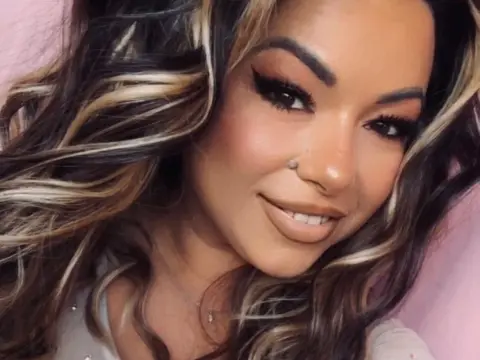 kitttty4play — Smiling woman with wavy highlighted hair and glamorous makeup in a soft pink setting.