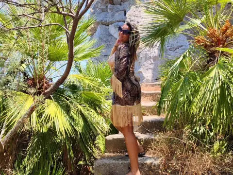 kitttty4play — Stylish woman in a patterned fringe dress posing amidst lush tropical palm trees on a sunny rocky hillside.