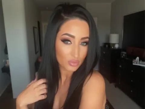 Glamorous woman with long dark hair and smoky eye makeup posing in a modern bedroom setting.