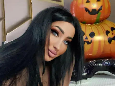 Woman with long black hair and glamorous makeup posing near festive orange Halloween pumpkin balloons.