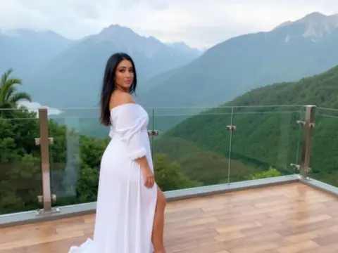 Woman in elegant white maxi dress posing on a balcony overlooking lush green mountains under a cloudy sky.