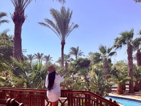 Woman in a white summer dress standing on a wooden bridge overlooking a tropical palm tree resort garden.