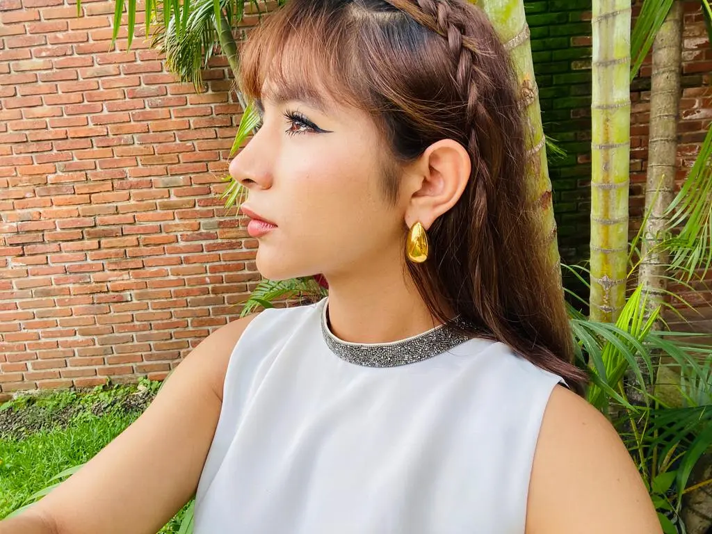 NatalyArango — NatalyArango poses in a white sleeveless top with a silver collar, showing her long brown hair in a side braid against a brick wall and palm trees.