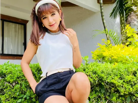 NatalyArango — NatalyArango poses outdoors in a white sleeveless top and black shorts, wearing a white headband and gold earrings while sitting near green bushes.