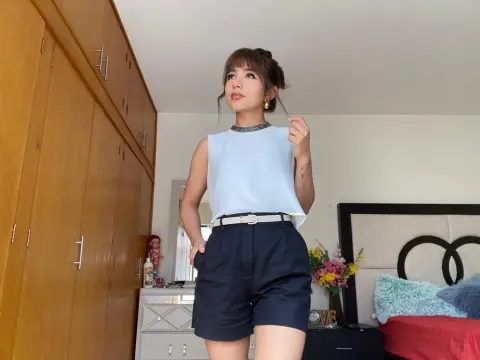 NatalyArango — NatalyArango poses in a white sleeveless top and dark blue shorts with a white belt, standing in a bedroom near a wooden wardrobe.