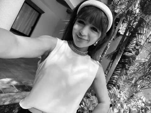 NatalyArango — NatalyArango smiles wearing a white sleeveless top and a thick white headband, posing outdoors near palm leaves in a black and white photo.