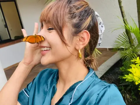 NatalyArango — NatalyArango smiles while a large orange butterfly rests on her nose, wearing a teal satin robe and gold earrings outdoors near green plants.