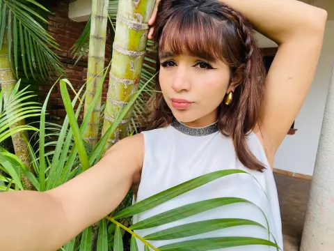 NatalyArango — NatalyArango poses outdoors near palm leaves, wearing a white sleeveless top and a silver choker, with one arm raised behind her head.