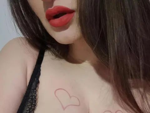 Annastea21x — Close-up of a woman with red lipstick and a heart tattoo wearing a black lace camisole in soft lighting.