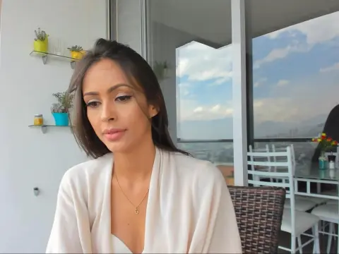Young woman in a white cardigan sitting on a bright balcony with a scenic city view.