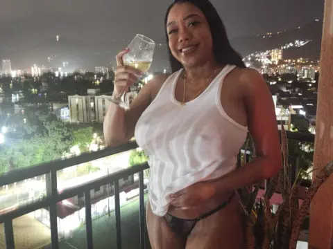 Tyfaniebony — Smiling woman in a white tank top holding a glass of wine on a balcony overlooking a city at night.