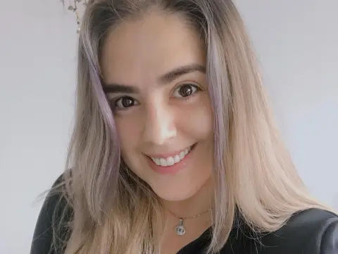 PamelaFI0RI — PamelaFI0RI smiles with long, light blonde hair and a silver pendant necklace, wearing a black top against a plain white wall.