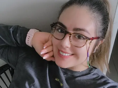 PamelaFI0RI — PamelaFI0RI smiles wearing glasses and a grey long-sleeve shirt, with her hair pulled back in a ponytail and a pink watch on her wrist.