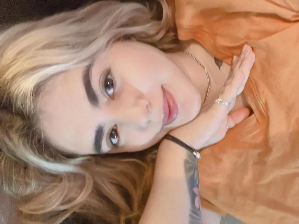 Blonde woman in an orange top relaxing indoors with a thoughtful expression.