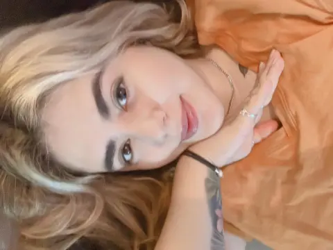 PamelaFI0RI — Blonde woman in an orange top relaxing indoors with a thoughtful expression.