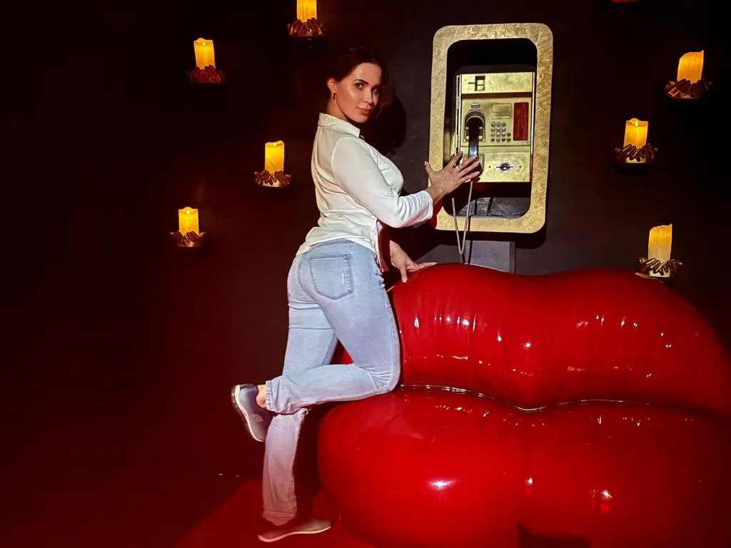 AdultsShowN1 — Woman in white shirt and jeans sitting on a red sofa in a moody, candlelit room near a vintage phone.