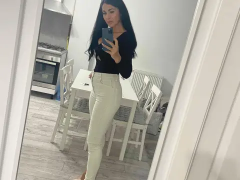 ELENA001 stands in a kitchen wearing a black long-sleeve top and tight white jeans.