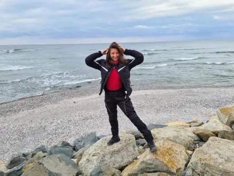 Smiling woman in a black jacket and red top standing on rocks by the ocean under a cloudy sky.