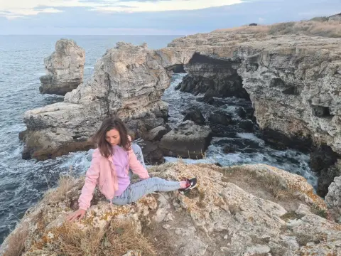 Young woman in a pink jacket and jeans sitting on coastal rocks by the ocean at sunset.