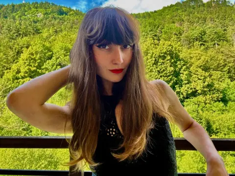 Woman with long brown hair and red lipstick posing on a balcony against a lush green forest background.