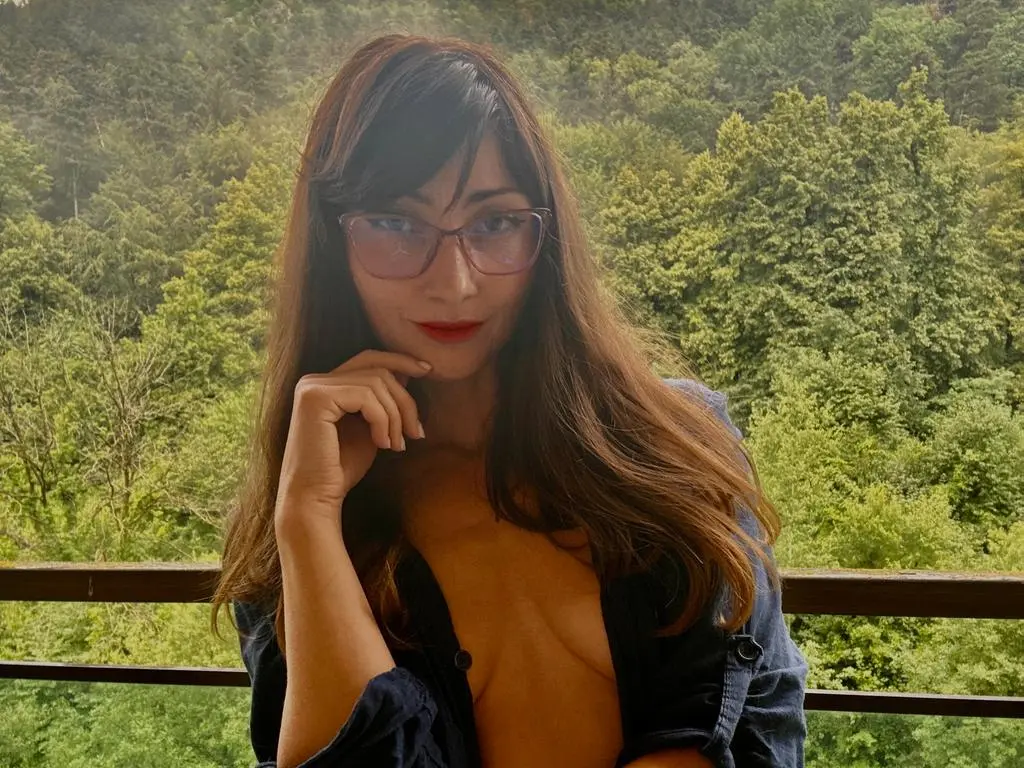 Smiling woman with long brown hair in a dark shirt relaxing on a balcony overlooking a lush green forest.