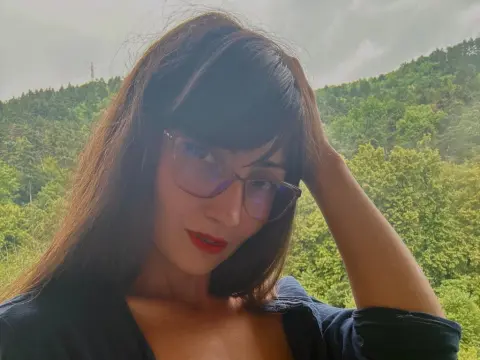 Young woman with glasses and red lipstick posing against a lush green forest background.