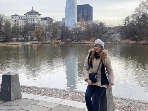 Woman in a beige winter coat and black scarf standing by a city lake on a cloudy day.