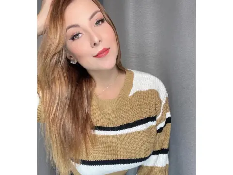 Smiling woman with long blonde hair wearing a tan and white striped knit sweater in a bright indoor setting.