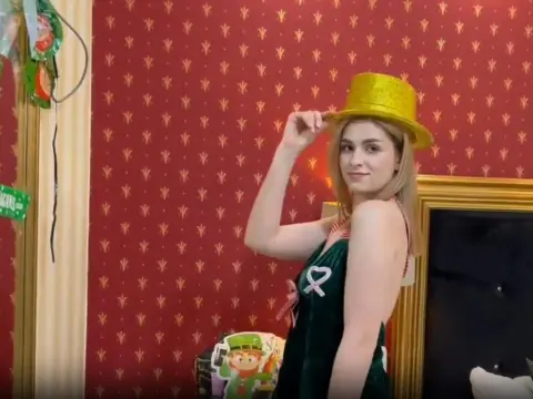 Smiling woman in a green velvet dress and gold top hat posing against a red patterned wallpaper background.