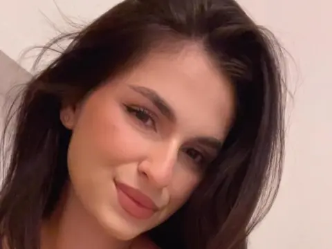 Smiling young woman with long dark hair and natural makeup in a soft indoor setting.