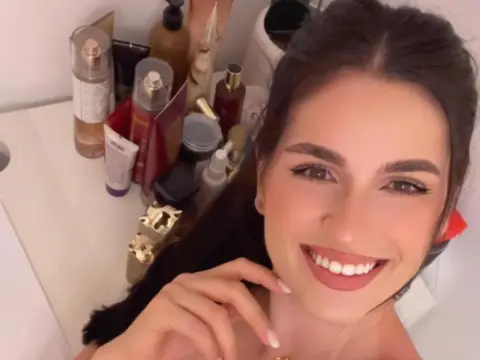 Smiling young woman with long dark hair posing in a bright room surrounded by various beauty and skincare products.