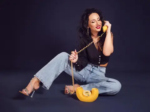 Stylish woman in denim jeans and silver heels talking on a vintage yellow telephone against a dark background.