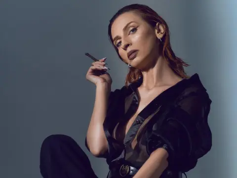 MinnieDrew — Elegant woman in a black sheer blouse smoking a cigarette in a moody studio setting.